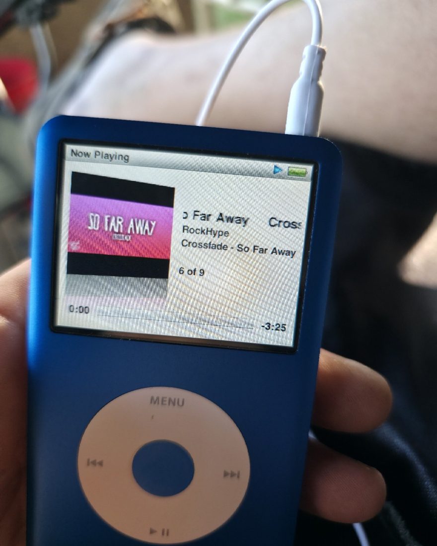 A blue iPod Classic held in a hand, earbuds plugged in, a song on screen.
