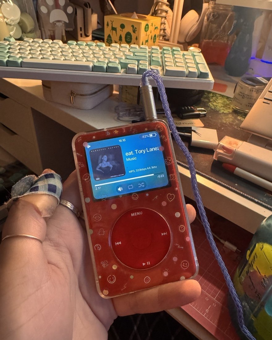 A red translucent iPod Classic held in a hand at a desk, a track playing on screen.